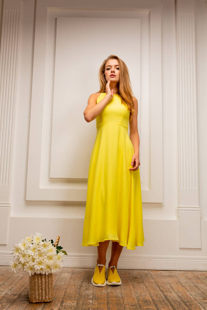 Sarı elbiseli, sarı ayakkabılı, mavi stüdyo arka planında çiçekli güzel sarışın kız. - Fotoğraf, Görsel