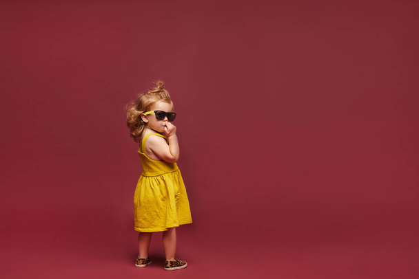 Cool stylish little girl in yellow dress and sunglasses and sunglasses isolated at the pink background. Child fashion. Copy space - Valokuva, kuva