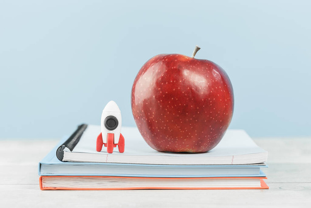 Organized office or school desk. Toy rocket and big red apple on pile of notebooks on wooden desk. Copy space for back to school or education knowledge craft and innovative startup concept - Photo, Image