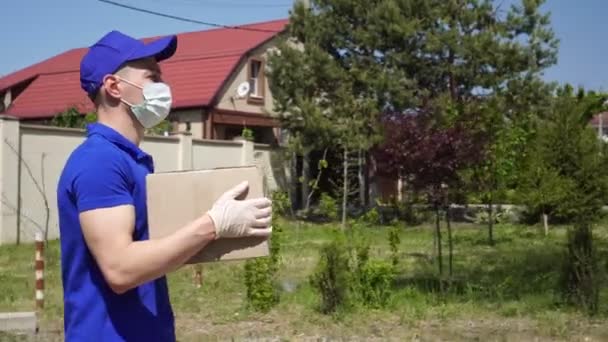 A delivery man in a medical mask and gloves carries a cardboard box - Footage, Video