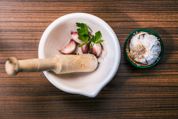 Ετοιμάζοντας μια συνταγή για να φτιάξετε αϊόλι με παραδοσιακό και σπιτικό τρόπο. Συστατικά και σκεύη για την προετοιμασία του σκόρδου πετρελαίου. Διαφορετικές σκελίδες σκόρδου, φύλλα μαϊντανού, κεφαλή σκόρδου και ένα κονίαμα στην κορυφή ενός ξύλινου πάγκου vintage. - Φωτογραφία, εικόνα