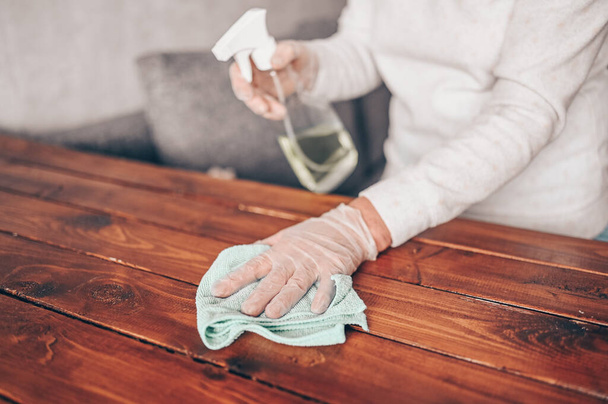 Großaufnahme der Reinigung des heimischen Holztisches, Desinfektion der Küchentischoberfläche mit einer antibakteriellen Sprühflasche, Waschen der Oberflächen mit Handtuch und Handschuhen. COVID-19 Prävention im Inneren. - Foto, Bild