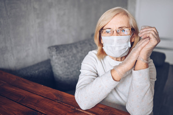 Lonely depressed sad older elderly senior woman in face protective mask, glasses, and medical gloves sitting at home self isolation quarantine during coronavirus COVID19 pandemic. Stay home concept - Photo, Image