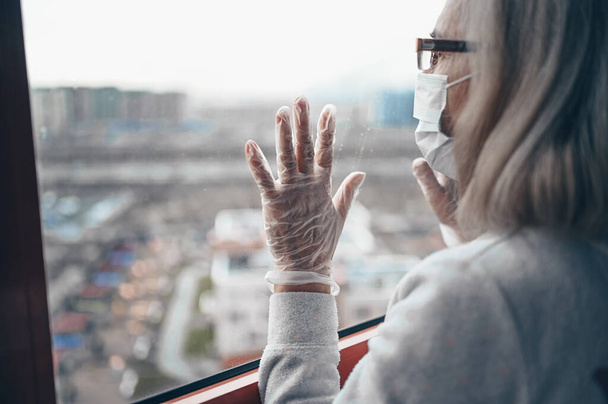Eenzame bejaarde droevige vrouw met een medisch masker en handschoenen die naar ramen kijkt, blijft thuis geïsoleerd voor zelfquarantaine. Concept home quarantaine, preventie COVID-19, Coronavirus uitbraak situatie - Foto, afbeelding