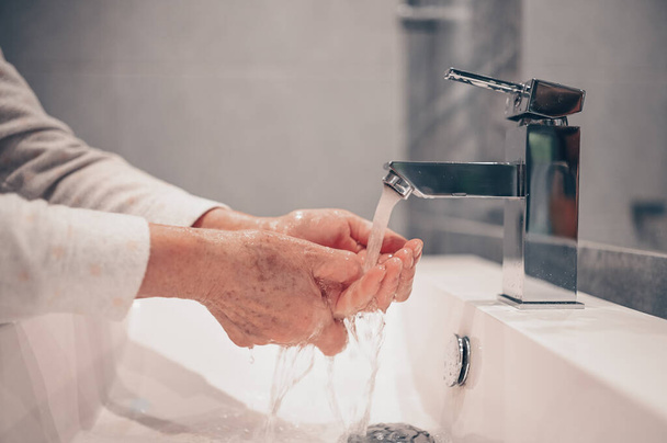 Lavage des mains mousser savon liquide frotter les poignets lavage des mains étape senior femme rinçant dans l'eau à l'évier du robinet de salle de bains. Lavez-vous les mains pour prévenir la propagation de COVID-19. Épidémie de coronavirus
. - Photo, image