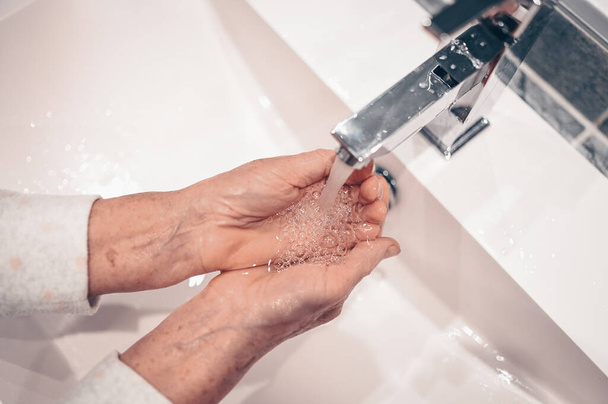 Lavage des mains mousser savon liquide frotter les poignets lavage des mains étape senior femme rinçant dans l'eau à l'évier du robinet de salle de bains. Lavez-vous les mains pour prévenir la propagation de COVID-19. Épidémie de coronavirus
. - Photo, image