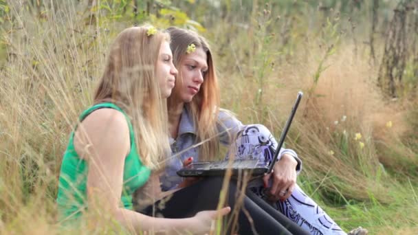 2 つの女性の友人は草の上に座っているとオンラインで話しているラップトップを使用してください。 - 映像、動画