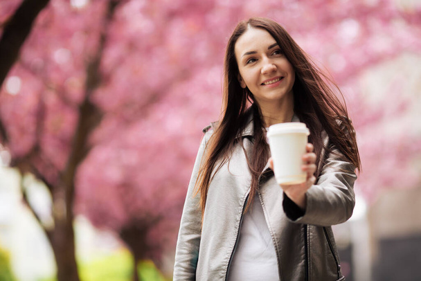 brunette girl in jacket and jeans in the spring outdoors in the city against the backdrop of flowering sakura trees posing - Fotografie, Obrázek