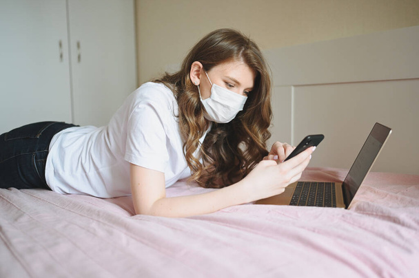Nuori eurooppalainen nainen kasvojen lääkekasvonaamiossa työskentelee kannettavan tietokoneen ja puhelimen sängyssä sepelvaltimoviruksen eristyksen aikana. Covid-19-pandemiavirus. Etäisyys verkossa työtä kotoa käsite
. - Valokuva, kuva