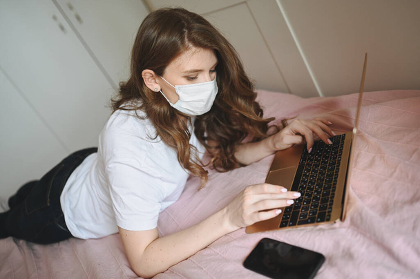 Młoda Europejka w masce medycyny twarzy pracująca na laptopie i telefonie w łóżku podczas kwarantanny domowej. Wirus Covid-19 pandemii Corony. Praca online na odległość od koncepcji domu. - Zdjęcie, obraz