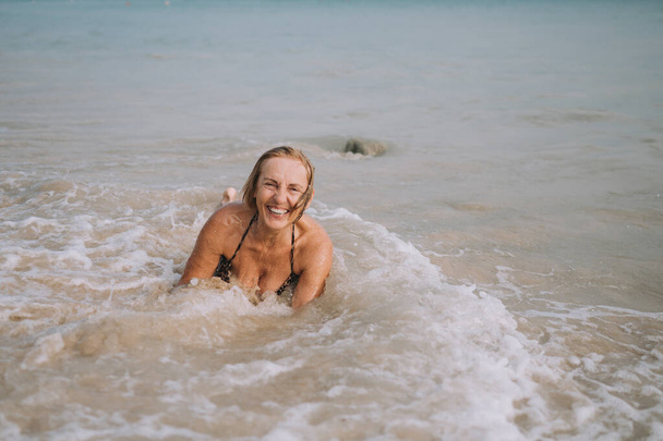 Χαρούμενη χαμογελαστή ηλικιωμένη ηλικιωμένη τουρίστρια που παίζει στο νερό και κολυμπά στα μεγάλα κύματα στην παραλία του ωκεανού. Ταξιδεύοντας κατά μήκος της Ασίας, την έννοια του ενεργού τρόπου ζωής. - Φωτογραφία, εικόνα