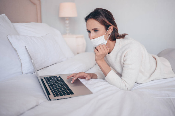 顔の薬マスクの若いヨーロッパの女性は、コロナウイルスの隔離家庭の隔離中に寝室のラップトップで働いています。Covid-19パンデミックコロナウイルス。ホームコンセプトからの距離オンライン作業. - 写真・画像