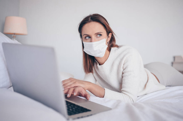 Young european woman in face medicine mask working on a laptop in bedroom during coronavirus isolation home quarantine. Covid-19 pandemic Corona virus. Distance online work from home concept. - Foto, Bild