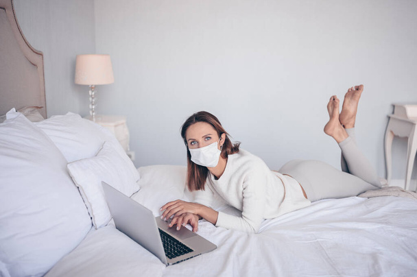 顔の薬マスクの若いヨーロッパの女性は、コロナウイルスの隔離家庭の隔離中に寝室のラップトップで働いています。Covid-19パンデミックコロナウイルス。ホームコンセプトからの距離オンライン作業. - 写真・画像