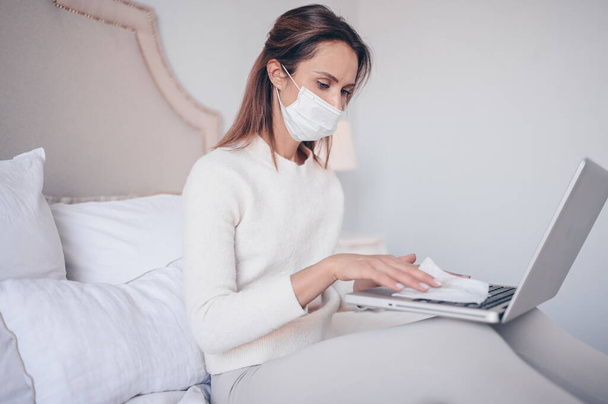 European woman in face mask in bedroom during coronavirus isolation home quarantine cleaning laptop by hand sanitizer, using cotton wool with alcohol to wipe to avoid contaminating with Corona virus. - Zdjęcie, obraz