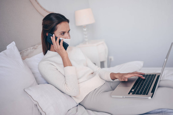 Jonge vrouw in bed, spreekt telefoon en werkt aan een laptop. Home quarantaine zelfisolatie tijdens pandemie Corona virus. Afstandswerk van thuis. COVID-19 concept om veilig thuis te blijven red levens - Foto, afbeelding