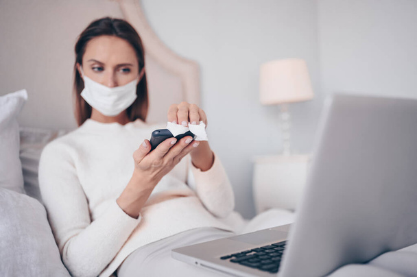 Mladá Evropanka s obličejovou maskou v ložnici s laptopem během izolace koronaviru domácí karanténa čištění telefonu dezinfekčním prostředkem na ruce, s použitím bavlněné vlny s alkoholem. Koronavirus COVID-19 pandemie - Fotografie, Obrázek