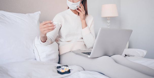 Αγνώριστη άρρωστη γυναίκα με μάσκα προστασίας προσώπου ξαπλωμένη στο κρεβάτι με φορητό υπολογιστή που κρατάει θερμόμετρο και χάπια στην απομόνωση καραντίνας στο σπίτι. Online δουλειά από το σπίτι. Ιογενής λοίμωξη του κερατοειδούς COVID-19 έννοια. - Φωτογραφία, εικόνα