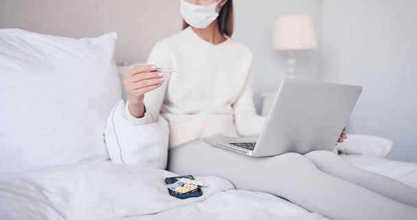 Unrecognizable sick woman in face protection mask lying in bed with laptop holding thermometer and pills at home quarantine isolation. Online work from home. Corona virus infection COVID-19 concept. - Foto, afbeelding