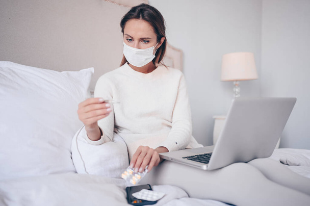Sick woman in face protection mask lying in bed holding thermometer and pills at home quarantine isolation. Corona virus COVID-19 concept. Suspecting Infection. Respiratory illness first symptom fever - Photo, Image