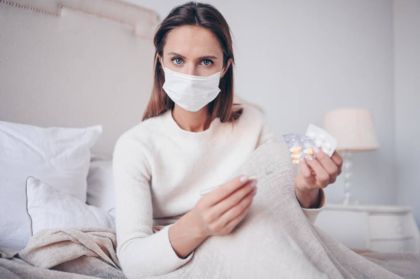 Nemocná žena v ochranné masce, ležící v posteli a držící teploměr a pilulky v domácí karanténě. Koncept Corona virus COVID-19. Podezření na infekci. Respirační onemocnění první symptom horečka - Fotografie, Obrázek