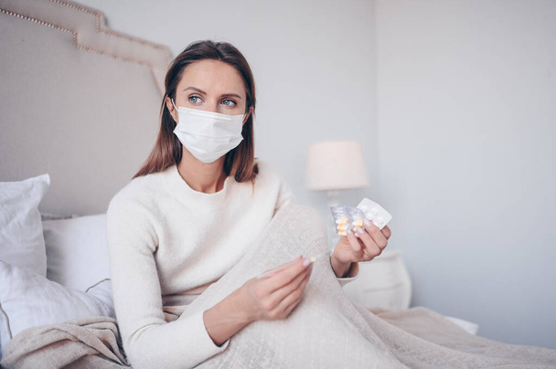 Chora kobieta w masce ochronnej leży w łóżku trzymając termometr i pigułki w izolatce domowej kwarantanny. Wirus korony COVID-19 koncepcja. Podejrzewam infekcję. Choroby układu oddechowego gorączka pierwszego objawu - Zdjęcie, obraz