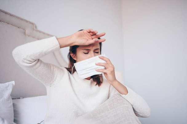 Objawy choroby koronawirusowej (COVID-19) - katar, ból gardła, kaszel, gorączka. Młoda kobieta z maską na twarzy chora na infekcję wirusową grypy rozprzestrzeniającą się w łóżku wirus korony w izolacji domowej kwarantanny - Zdjęcie, obraz