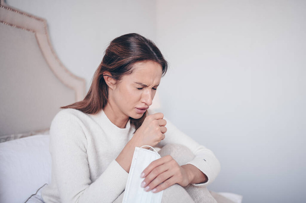 Симптомы коронавируса (COVID-19) - насморк, боль в горле, кашель, лихорадка. Молодая женщина в маске для лица, уставшая от вирусной инфекции гриппа, распространяющей коронный вирус, лежащий в постели в домашней карантинной изоляции
 - Фото, изображение