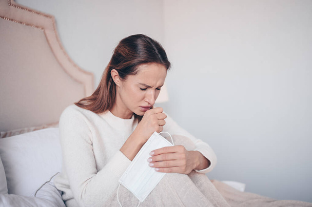 コロナウイルス病(COVID-19)症状-鼻水,喉の痛み,咳,発熱.インフルエンザウイルス感染症の顔のマスクを持つ若い女性が家の隔離隔離隔離でベッドに横たわっているコロナウイルスを広める - 写真・画像
