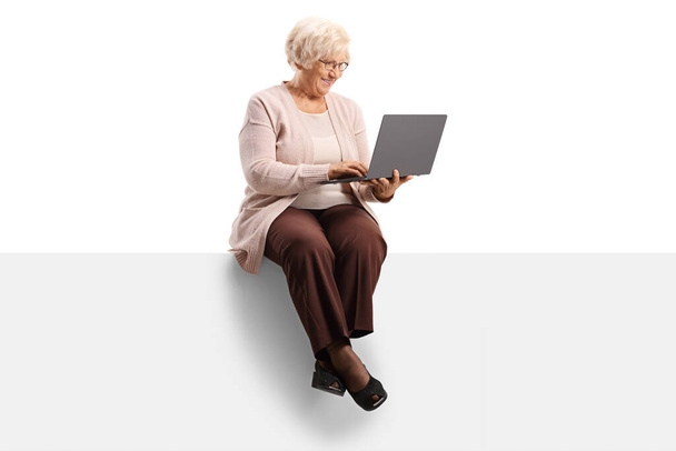 Cheerful elderly woman sitting on a blank panel and using a laptop computer isolated on white background - Foto, imagen