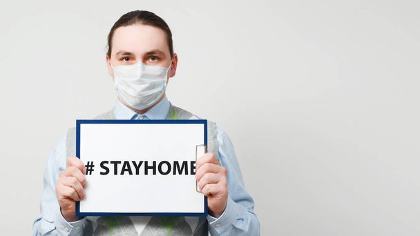 Male doctor in protective mask is holding paper with the hashtag stayhome on gray background. - Fotografie, Obrázek
