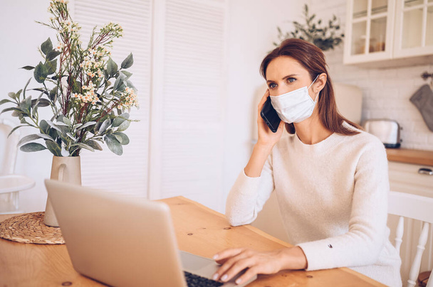 Egy európai beteg nő orvosi maszkban okostelefonnal beszél és egy laptopon dolgozik a konyhai karantén izolációjában Covid-19 pandémiás Corona vírus. Online munka, maradj otthon. Hőmérő és tabletták - Fotó, kép