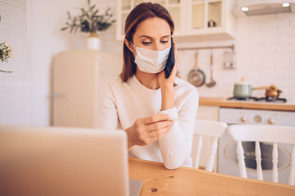 Europäische kranke Frau mit medizinischer Maske, Smartphone und Thermometer in der Hand, an einem Laptop arbeitend, Quarantäne-Isolation Covid-19 Coronavirus Pandemie. Online arbeiten, zu Hause bleiben - Foto, Bild