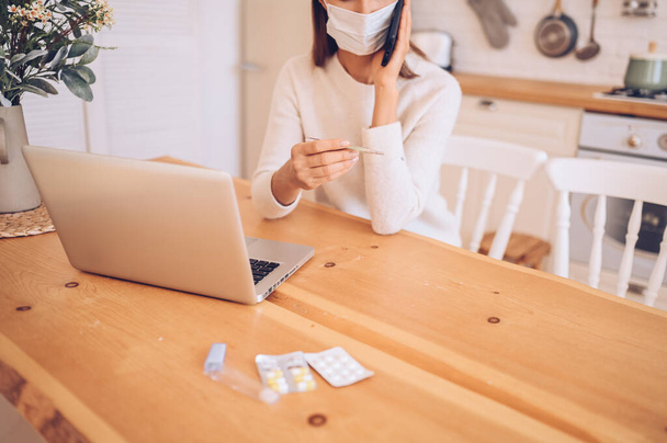 Nierozpoznana chora kobieta w masce medycznej mówiąca smartfonem, termometr w rękach, pracująca nad laptopem, izolacja domowa kuchni Covid-19 pandemia Wirus Corony. Praca online, pobyt w domu - Zdjęcie, obraz