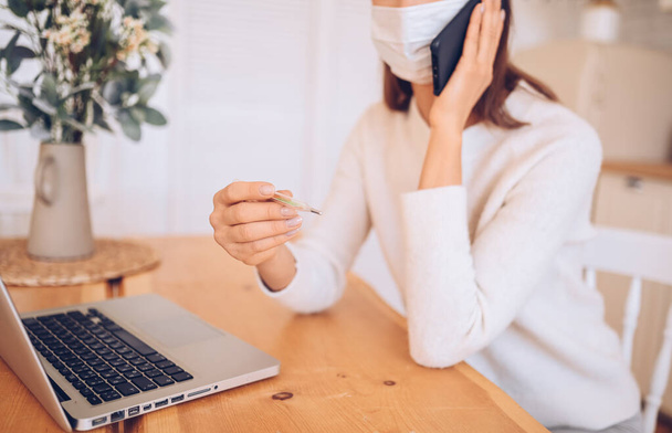 Unerkennbare kranke Frau in medizinischer Maske, Smartphone sprechend, Thermometer in den Händen, an einem Laptop arbeitend, Quarantäne-Isolation in der Küche, Covid-19 pandemische Coronavirus. Online arbeiten, zu Hause bleiben - Foto, Bild