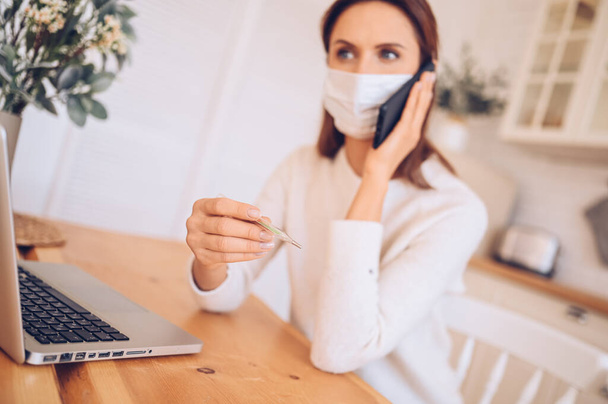 European sick woman in medical mask holding smartphone and thermometer in hands, working on a laptop, kitchen home quarantine isolation Covid-19 pandemic Corona virus. Distance online work, stay home - Photo, Image
