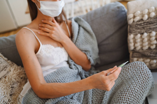 Sick unrecognizable woman in face protection mask on couch with grey comfortable blanket holding thermometer, home quarantine self isolation. Corona virus infection. COVID-19 concept promote stay safe home save lives - Fotó, kép