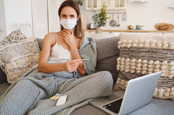 Sick young european woman in face protection mask on couch with grey comfortable blanket holding thermometer, home quarantine self isolation. Corona virus infection. COVID-19 concept promote stay safe home save lives - Photo, Image