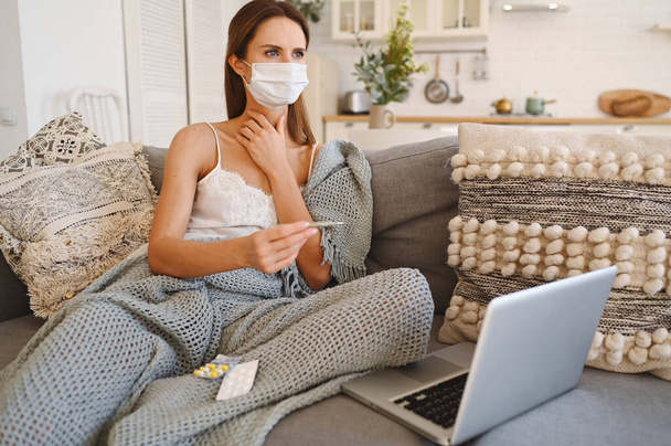 Хвора молода європейська жінка в масці для захисту обличчя на дивані з сірою зручною ковдрою, що тримає термометр, ізоляцію домашнього карантину. Корона вірусна інфекція. Концепція COVID-19 сприяння безпечному життю
 - Фото, зображення