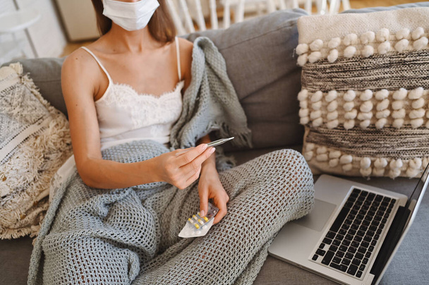 灰色の快適な毛布を保持温度計、自宅の隔離自己隔離とソファの上に顔の保護マスクで病気の認識できない女性。コロナウイルス感染症。COVID-19のコンセプトは、安全な家庭での生活を促進します。自宅からのリモートワーク - 写真・画像