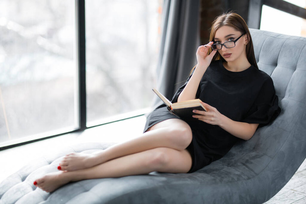 An American student in glasses and a black dress is lying at home on the sofa and reading a book on psychology. - Φωτογραφία, εικόνα