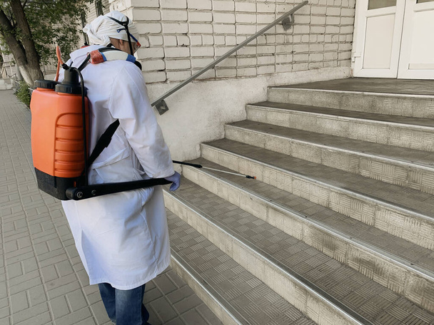 Ontsmetting door spray van het Corona-virus van de ingang van het woongebouw door de werknemer in speciaal uniform, epidemische preventie COVID-19 - Foto, afbeelding
