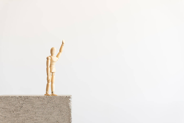 Wooden human figure standing isolated on a concrete block, waving with a raised hand. Symbol of self isolation and social distancing. - Photo, Image