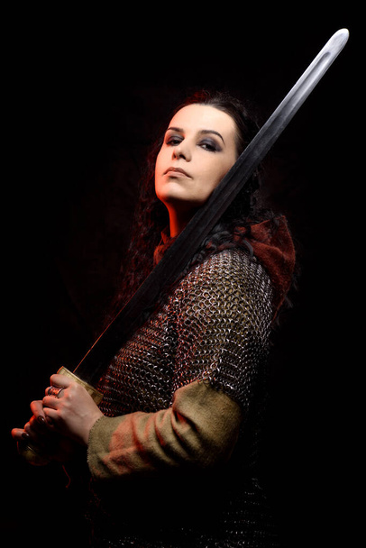 A dark-haired woman with a sword, a Scandinavian warrior in a chain mail against a dark background with red highlights - Foto, imagen