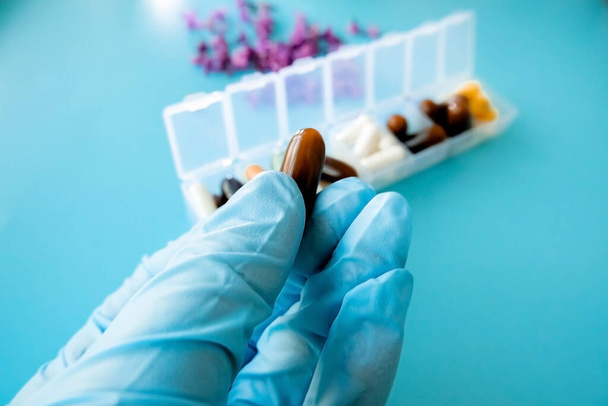 Una mano con un guante médico sostiene cápsulas multicolores, una caja de pastillas con medicamentos sobre un fondo azul
 - Foto, imagen