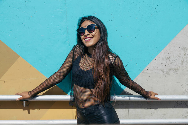 Bruna ragazza con i capelli lunghi e occhiali da sole in posa mentre è appoggiata a una ringhiera di fronte a una parete colorata e vestita in una maglia nera
 - Foto, immagini