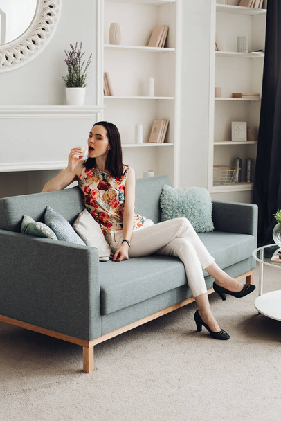 Stylish woman biting marshmallow on couch. casual clothing and heels sitting on modern couch with cushions and biting delicious marshmallow. - Photo, Image