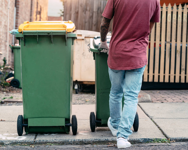 Een man is bezig met het opruimen van de prullenbak en de vuilnisbak op de stoep voordat hij door de vuilniswagen van de gemeente wordt opgehaald. Hij trekt aan de 120 & 240L vuilnisbakken. Concept: Afvalinzameling - Foto, afbeelding