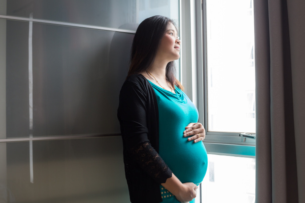Азійські вагітні жінки стоять біля вікна зі світлом, очікуванням концепції. - Фото, зображення