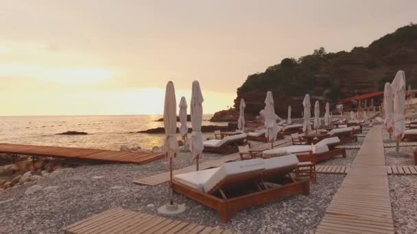 Flying the camera over the double sunbeds on rocky beach. Loungers for two with a soft mattress and a white umbrella. A wooden walkway on a pebble beach. Adriatic Coast in Montenegro at sunset - Footage, Video